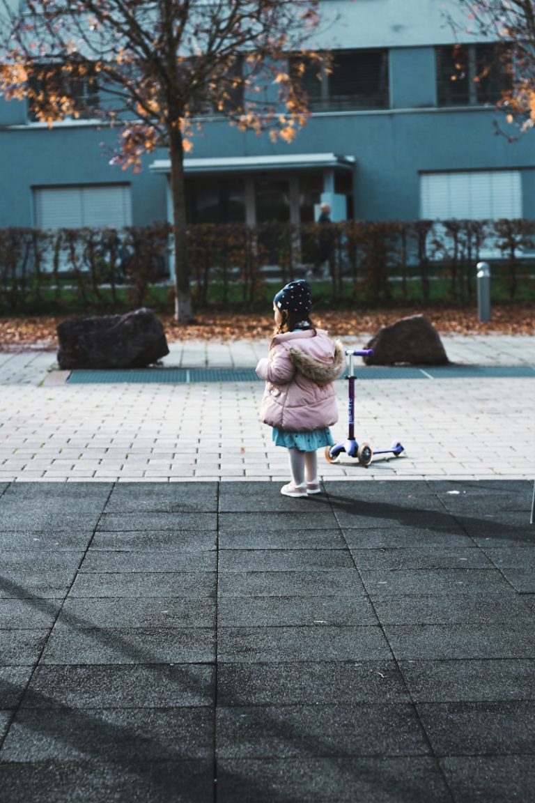girl in pink jacket riding kick scooter on gray concrete pavement during daytime